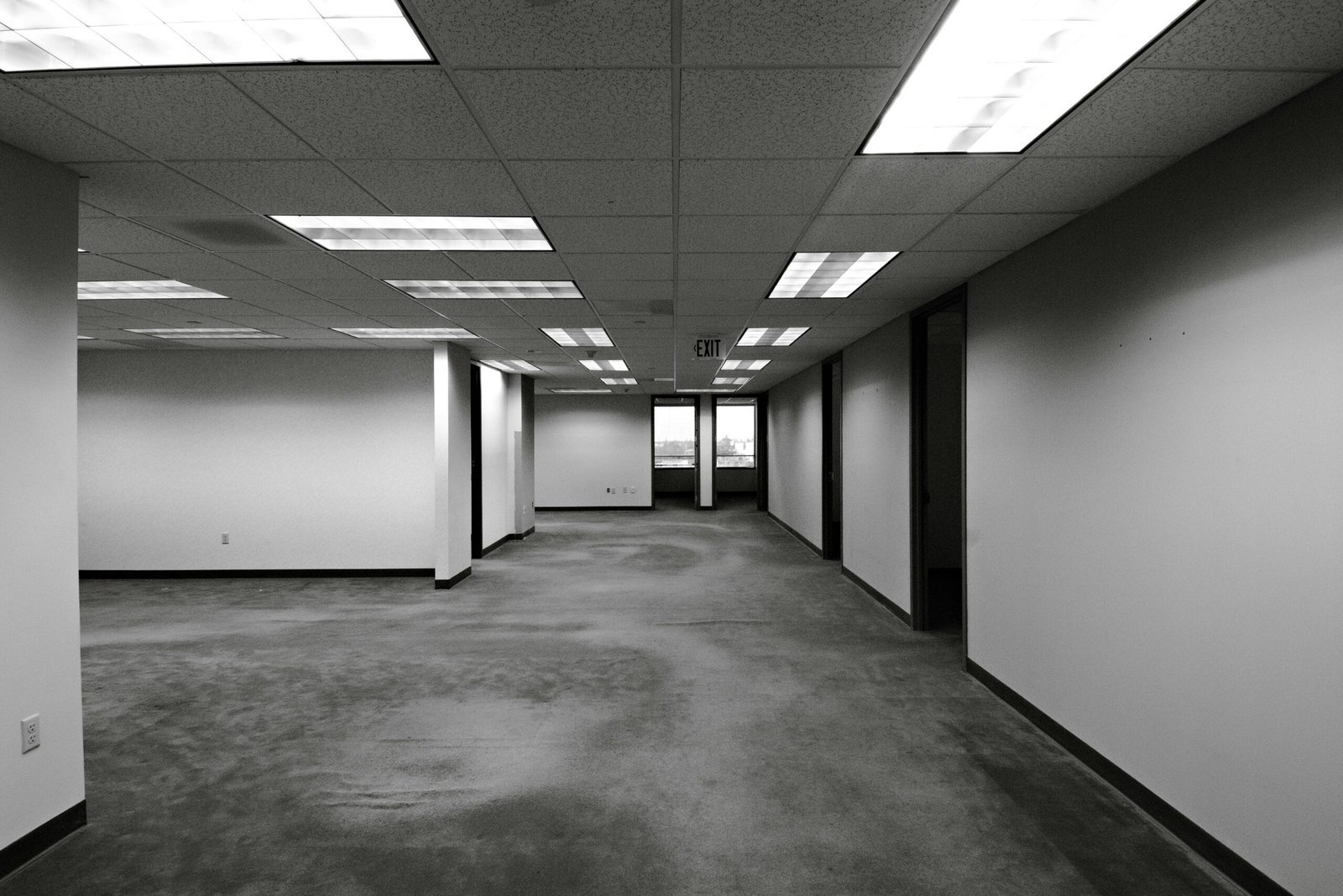 Black and white image of an empty office space with fluorescent lighting and exit sign.