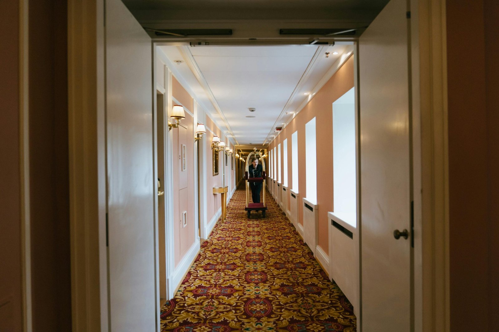 A bellboy pushes a luggage cart down a luxurious hotel corridor with elegant lighting and patterned carpet.